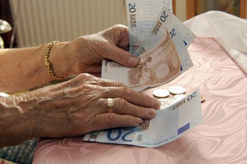 Pflegeheim-Kosten Händer einer alten Frau, die an einem Tisch sitzt, eventuell in einem Pflegeheim. Sie hält Geldscheine, auf dem Tisch liegen Münzen.