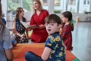 Das Bild zeigt eine Schulklasse mit mehreren Kindern und einer Lehrerin, die aus einem Buch vorliest. Im Vordergrund sieht man einen Jungen mit Down-Syndrom, der sich umdreht und direkt in die Kamera schaut. 