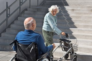 Ein älterer Mann im Rollstuhl und eine ältere Frau mit Rollator stehen am Fuß einer unüberwindbaren Treppe.