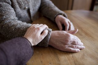 Ein alter Mann sitzt an einem Tisch, seine Hand liegt auf der Tischplatte. Eine jüngere Person hat liebevoll ihre Hand auf seinen Arm gelegt.