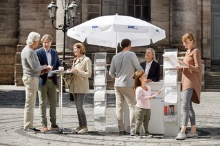 Ein VdK-Infostand in einer Fußgängerzone, eine Frau spricht mit einem Passanten, am Stehtisch daneben unterhalten sich Menschen. 