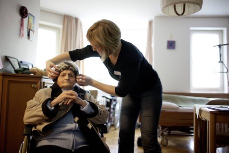 Eine alte Dame im Rollstuhl in ihrem Pflegeheim-Zimmer, eine jüngere Altenpflegerin hilft ihr gerade, ihre Mütze aufzusetzen.