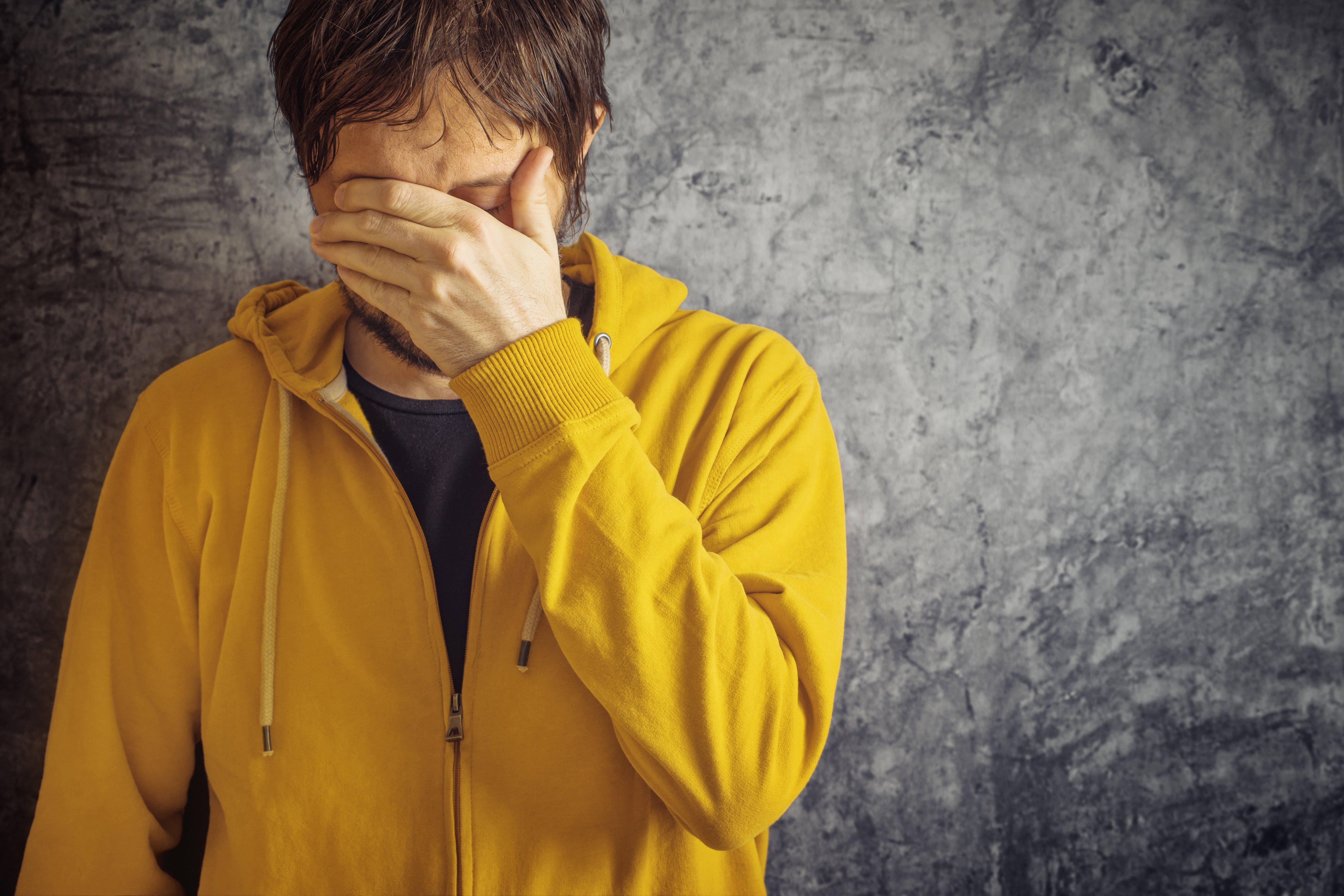 Ein Mann in gelber Sweatjacke steht vor einer grauen Wand, er h&auml;lt sich eine Hand vor die Augen, senkt den Kopf