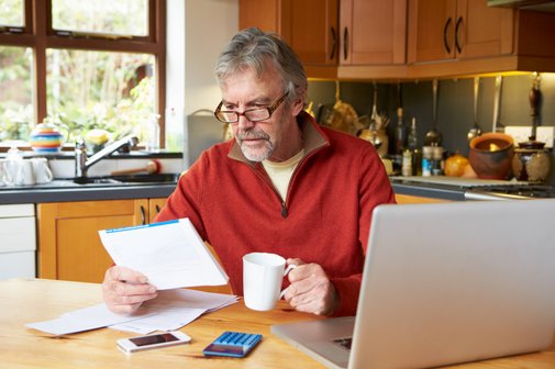 Älterer Mann sitzt in seiner Küche am Tisch, hat eine Kaffeetasse in der Hand und schaut Unterlagen an. Auf dem Tisch steht ein Notebook und ein Taschenrechner liegt vor dem Mann. 