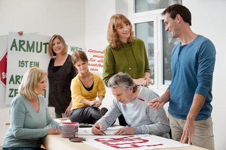 Eine Gruppe unterschiedlicher Menschen - M&auml;nner, Frauen, Alte und Junge, mit und ohne Behinderung, malt gemeinsam Demo-Plakate.