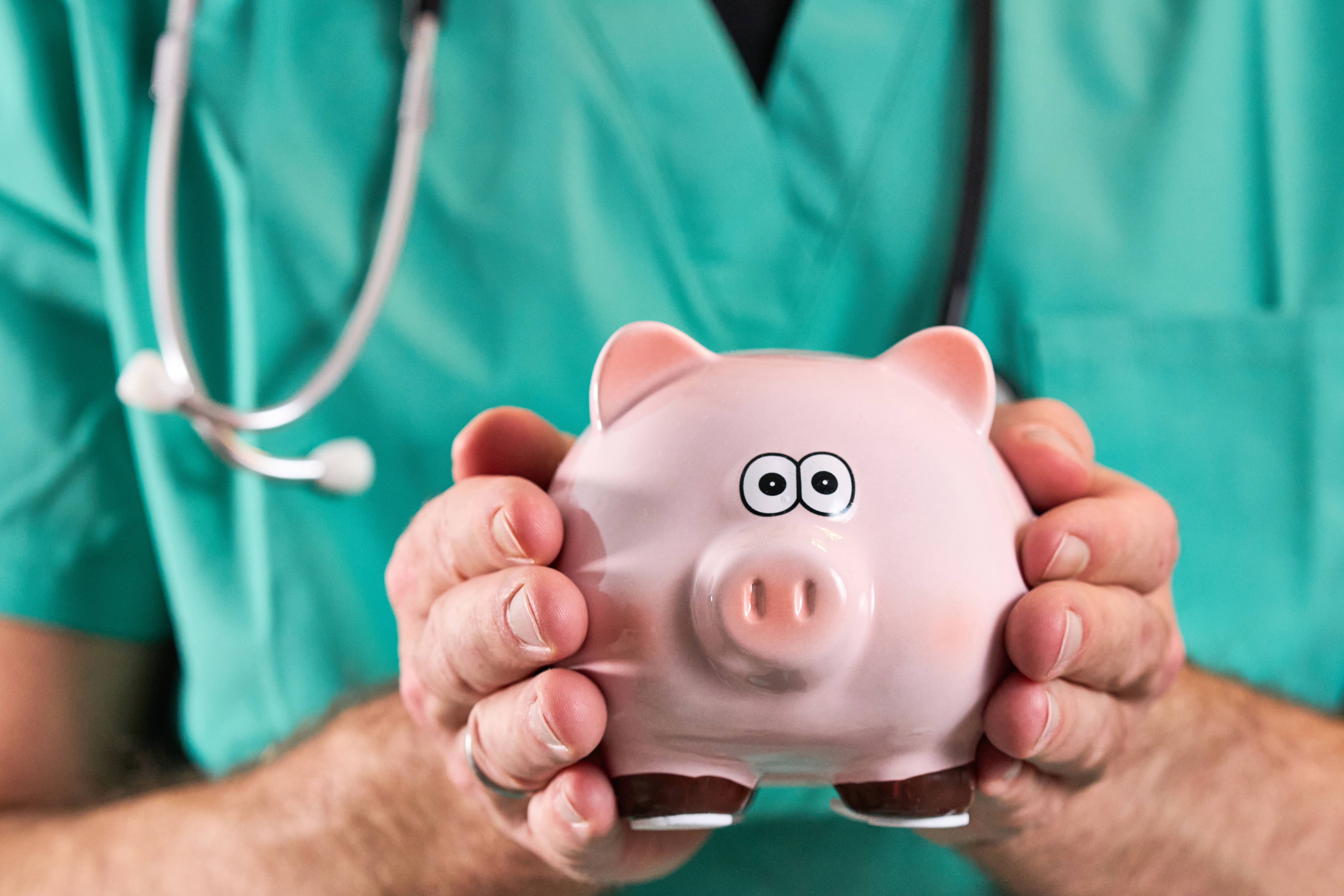 Kosten im Gesundheitssystem Ein Mann im grünen Arztkittel hält ein Sparschwein in beiden Händen, um den Hals hat er ein Stethoskop.