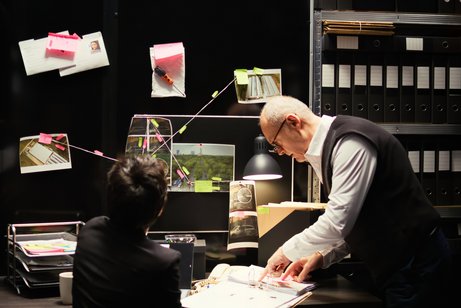 Zweo Detektive sitzen an einem Schreibtisch und schauen in ihre Unterlagen