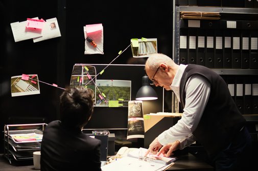 Zweo Detektive sitzen an einem Schreibtisch und schauen in ihre Unterlagen