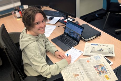 Eine junge Frau sitzt an einem Schreibtisch vor einem Laptop und einer aufgeschlagenen Zeitung.