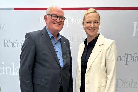 Ein Mann mit Brille und im Anzug steht neben einer blonden Frau mit weißem Sakko vor einem grauen Hintergrund.