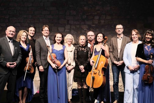 Frauen und M&auml;nner in festlicher Kleidung und mit Musikinstrumenten auf einer B&uuml;hne,