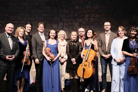 Frauen und M&auml;nner in festlicher Kleidung und mit Musikinstrumenten auf einer B&uuml;hne,