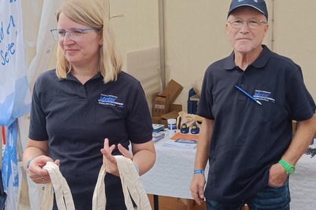 Eine Frau und ein Mann in blauen Polohemden verschenken Stofftaschen.