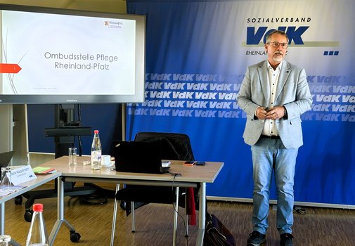 Ein Mann im Anzug steht vor einem blauen Hintergrund und einem Bildschirm mit einer Präsentation.