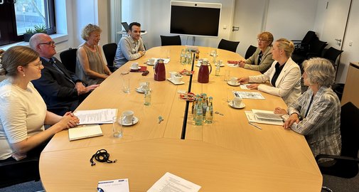 Sieben Frauen und Männer sitzen sich an einem großen ovalen Holztisch gegenüber.