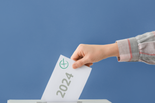 Ein Zettel mit 2024 als Aufschrift wird von einer Hand in eine Wahlurne geworfen.