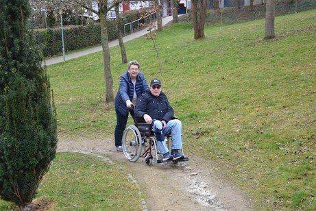 Eine Frau schiebt einen Mann im Rollstuhl über einen matschigen Weg.