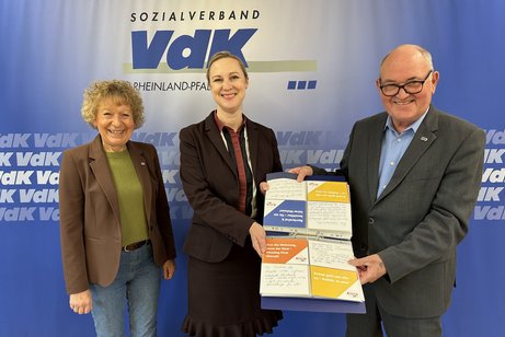 drei Menschen stehen vor einer VdK Wand und halten eine Mappe mit Postkarten in der Hand.