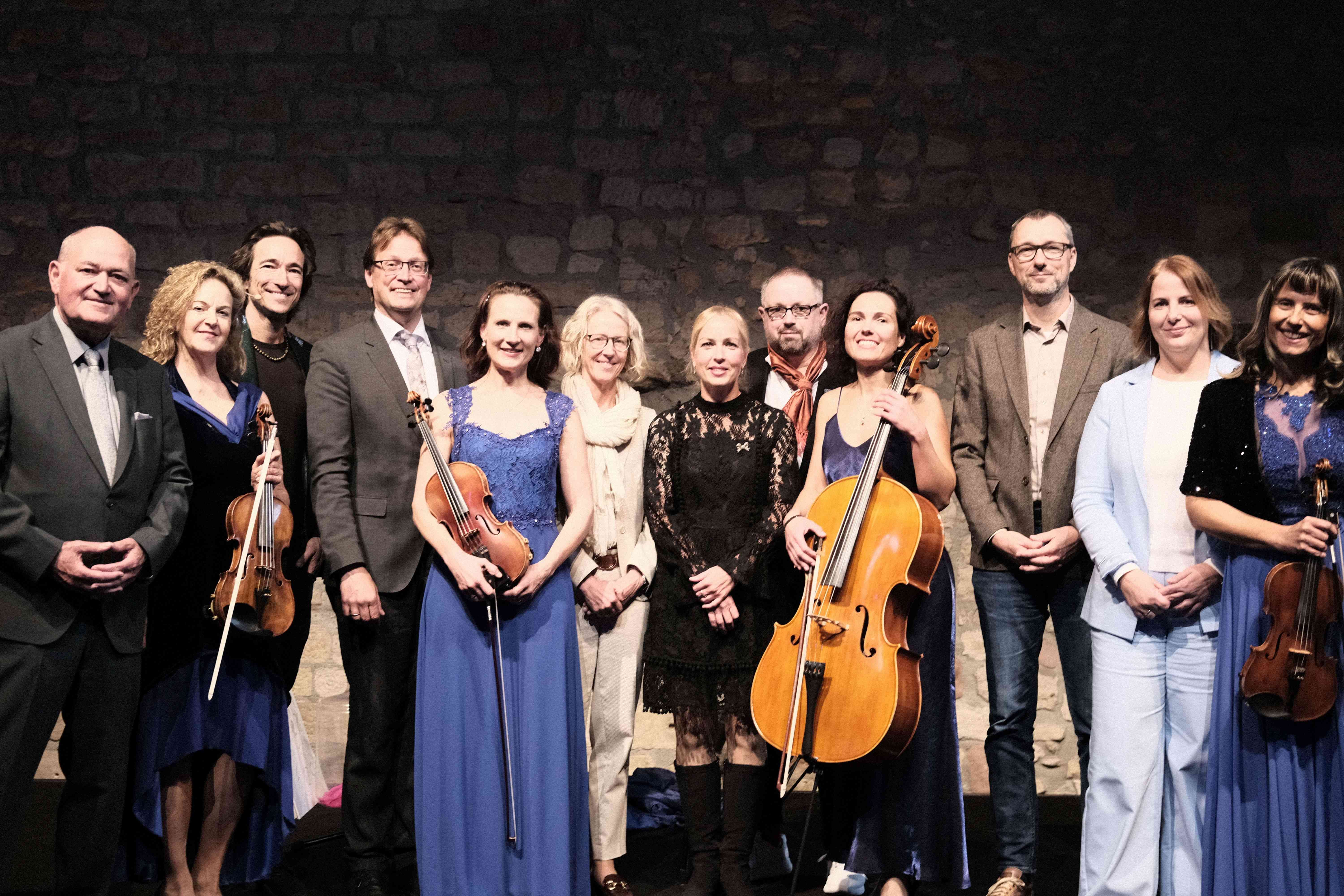 Frauen und M&auml;nner in festlicher Kleidung und mit Musikinstrumenten auf einer B&uuml;hne,