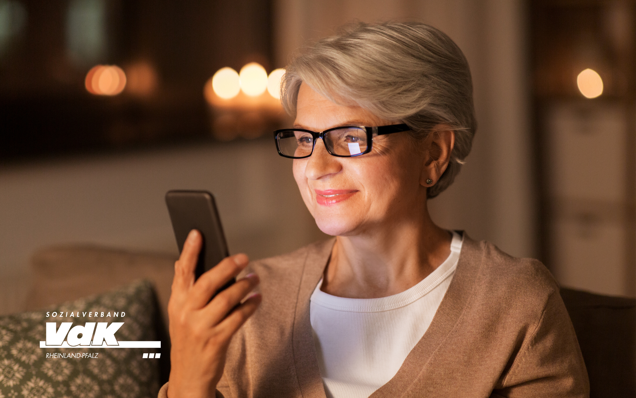 Ältere Frau mit Brille sitzt auf dem Sofa und schaut zufrieden auf ihr Smartphone.