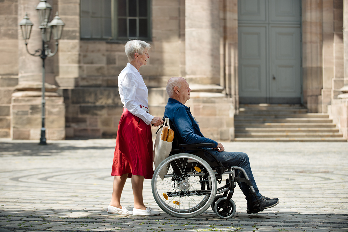 Frau schiebt ihren Mann Eine Seniorin schiebt ihren Mann, der im Rollstuhl sitzt, über einen Platz