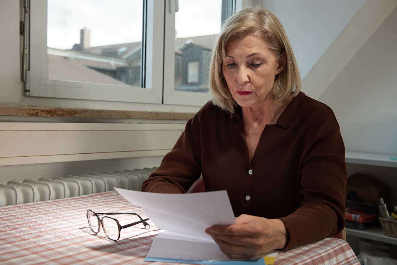 Eine &auml;ltere Frau sitzt an einem K&uuml;chentisch und liest einen Brief, sie sieht ernst und besorgt aus. 
