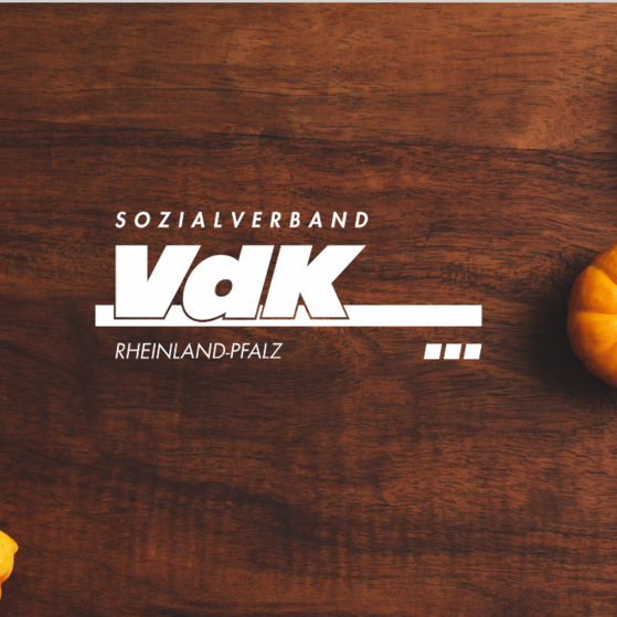 Weißes VdK-Logo vor braunem Hintergrund, umrahmt von Kürbissen.