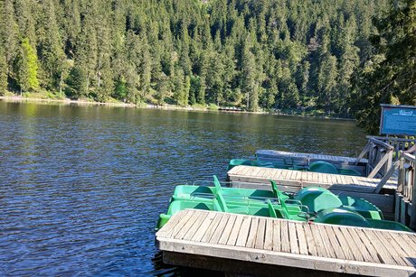 Zu sehen ist ein Blick über den Mummelsee und rechts im Bild liegen am Steg mehrere Tretboote.