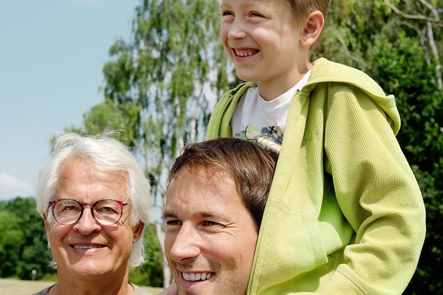 Männer aus drei Generationen Drei Männer - ein älterer Mann lächelt in die Kamera, ein jüngerer Mann blickt in die Ferne, auf seinen Schultern sitzt ein Junge.