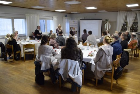 Menschen an runden Tischen, der Raum im &Uuml;berblick von hinten mit Leinwand