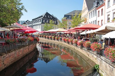 Innenstadtbereich von Saarburg am blumengeschm&uuml;ckten Ufer der Saar mit Tischen unter Sonnenschirmen