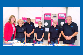 Eine Frau mit roter Jacke und 5 Personen in blauen VdK-T-Shirts stehen hinter einem Tisch.