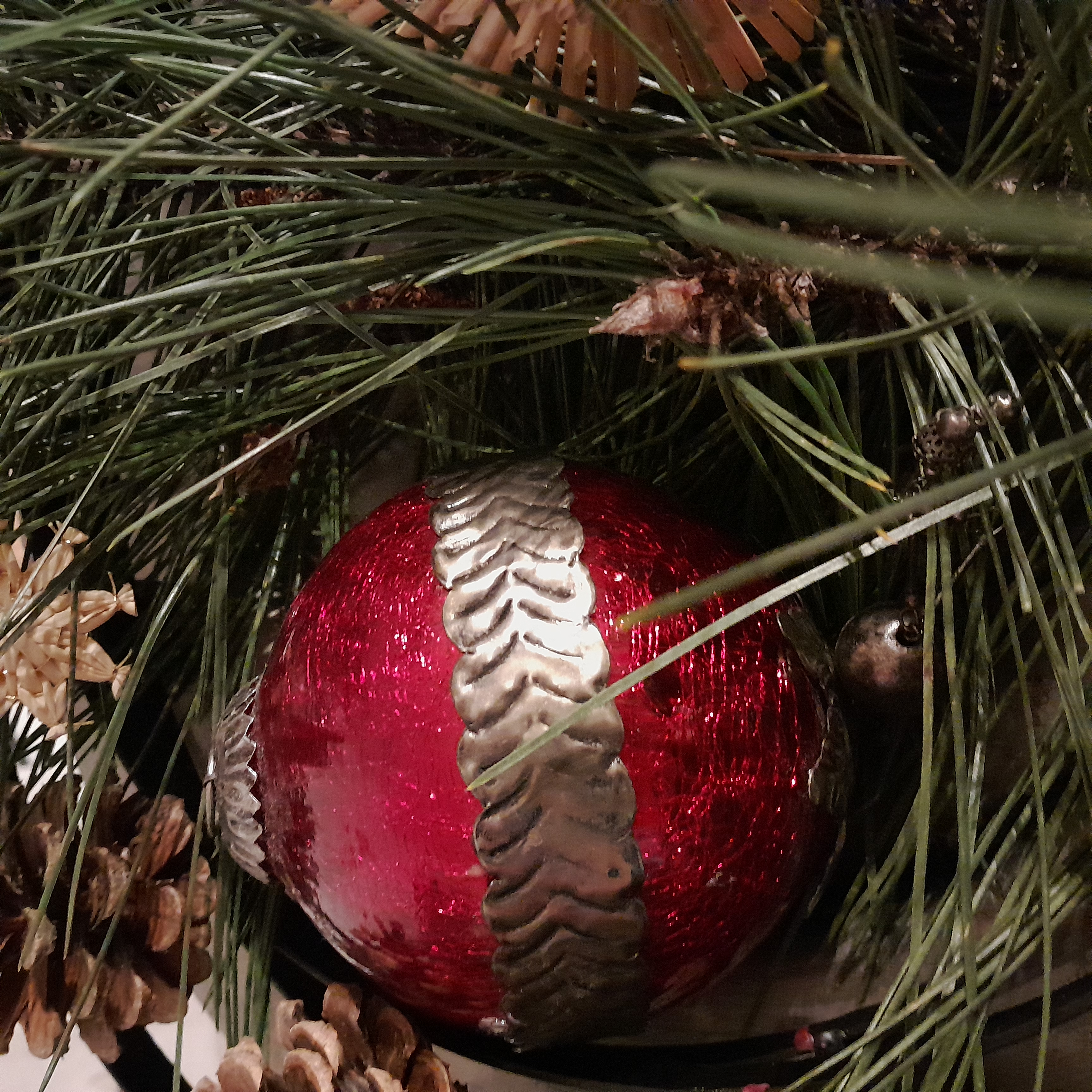 Dunkelrote Weihnachtskugel eingebettet in Kiefernnadeln, an der Seite sind Kiefernzapfen und Strohsterne